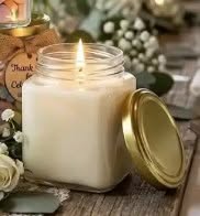 A white soy wax candle in a clear glass jar with a gold metal lid, shown lit on a rustic wooden surface with decorative flowers and greenery in the background.