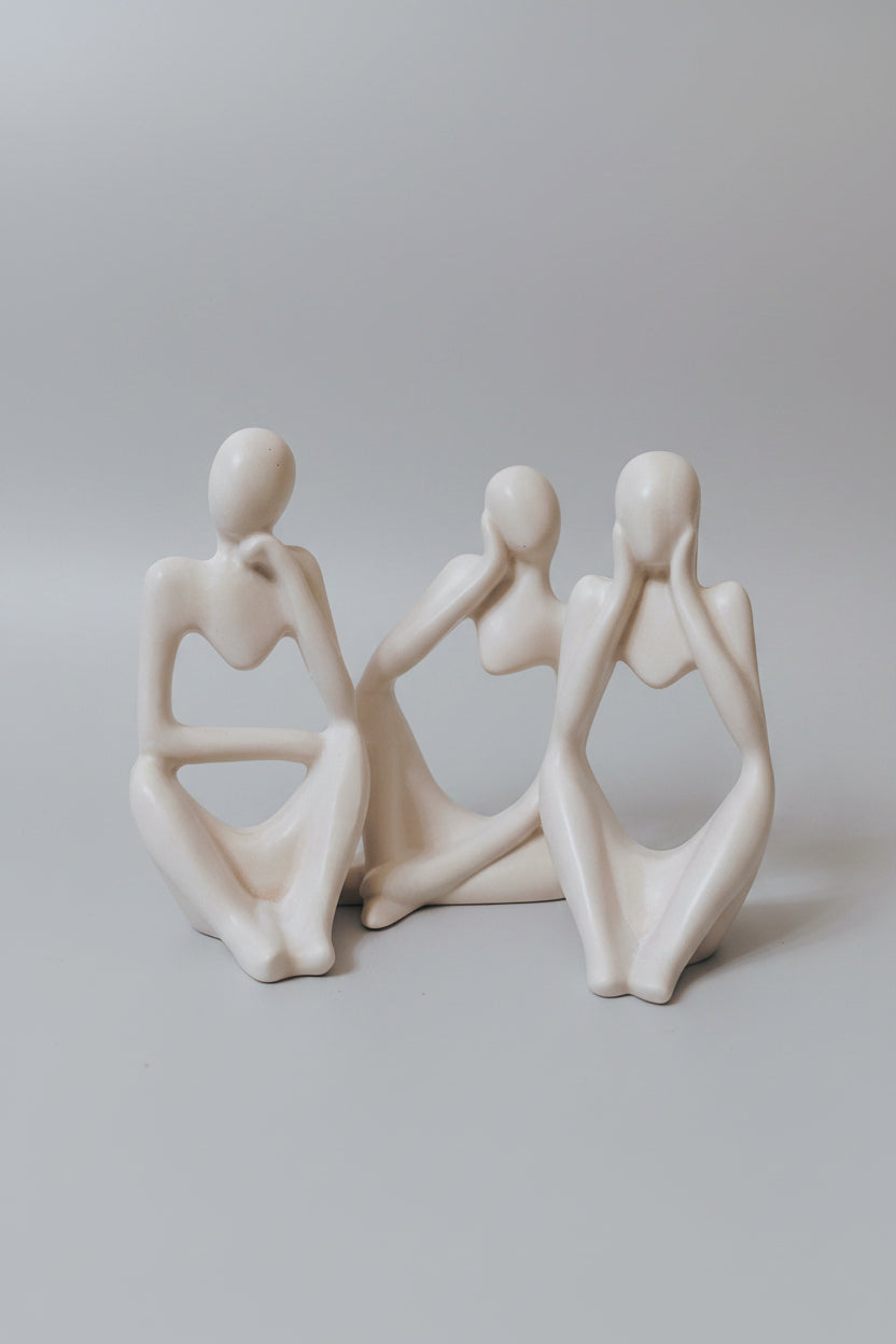 Three minimalist ceramic figurines of seated female figures in warm off-white, arranged side-by-side on a light surface.