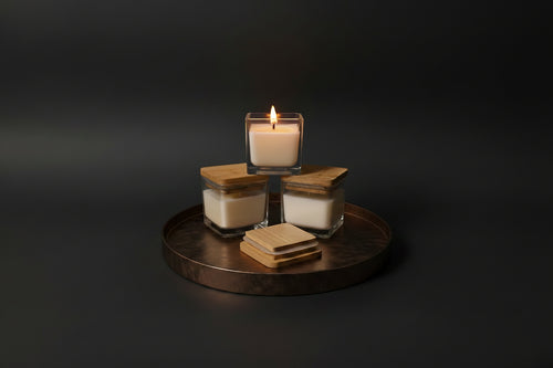 Three glass jar candles with light wood lids are arranged on a round white and green marble tray next to a matching square glass candle holder.