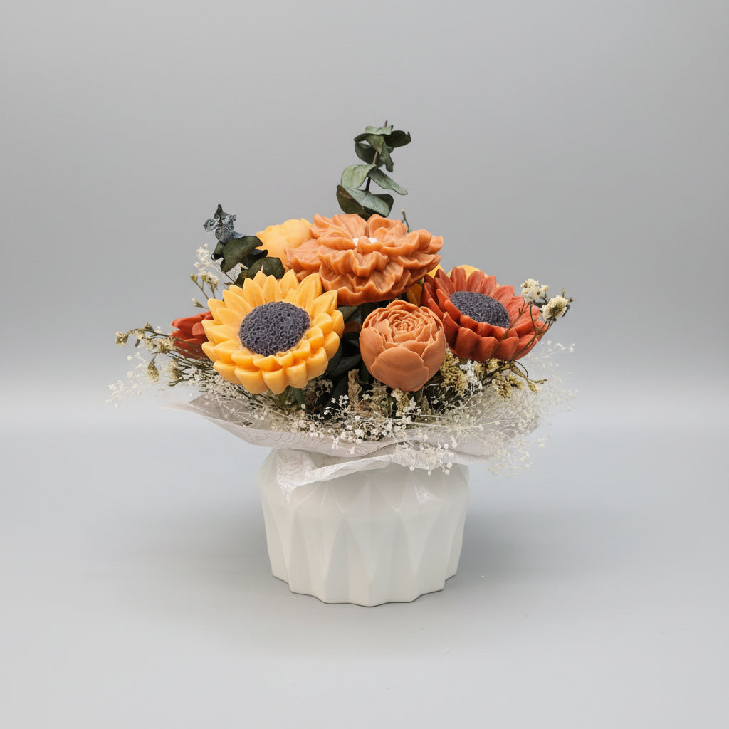 A decorative artificial flower arrangement in a faceted white pot, featuring orange and yellow sunflowers, peach roses, and dried green foliage.