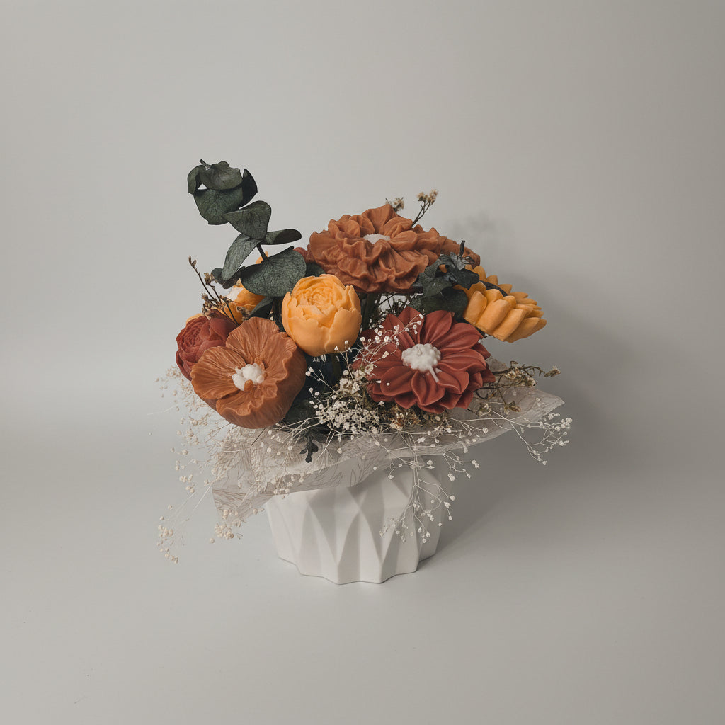 An artificial floral arrangement in a faceted white ceramic pot, featuring orange, yellow, and brown flowers with green and gray leaves, set against a plain beige wall and tabletop.