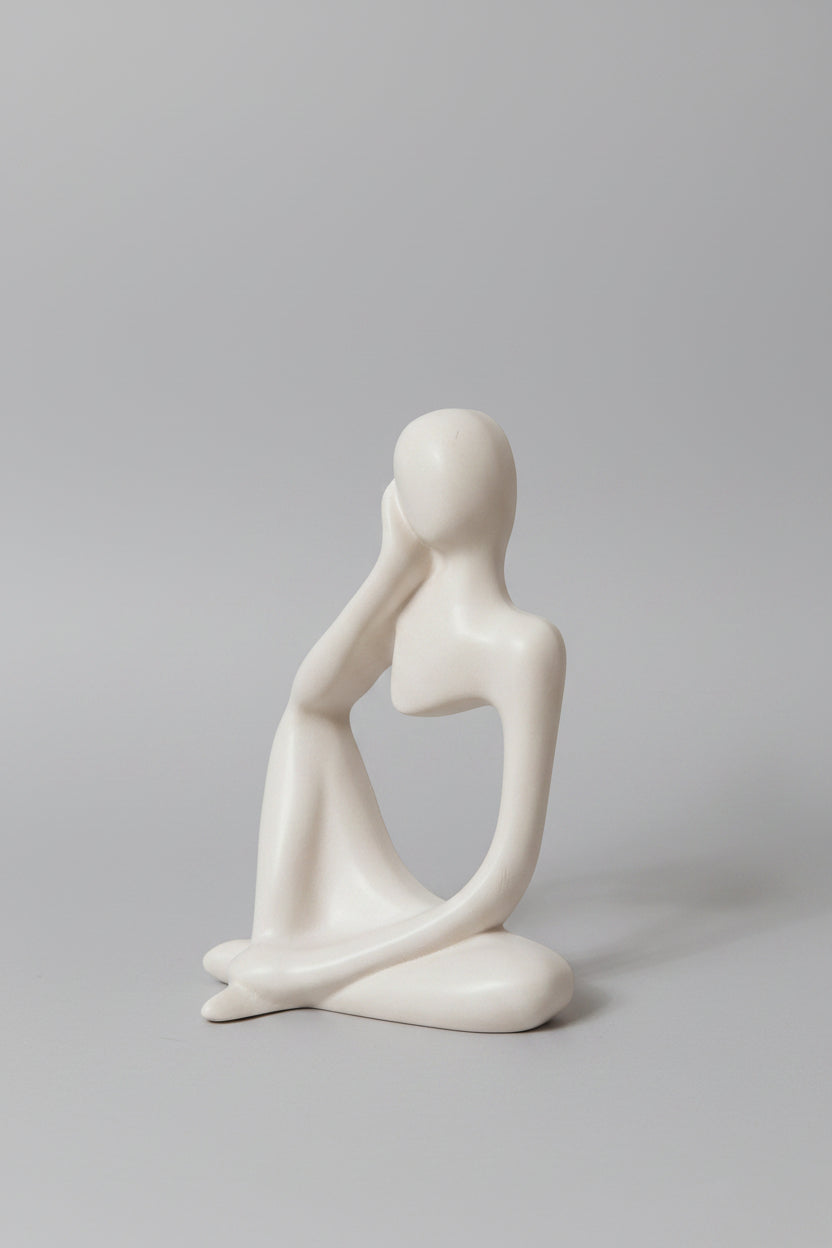 A minimalist white ceramic figurine of a seated, contemplative female figure with a blank face, resting on a light surface.