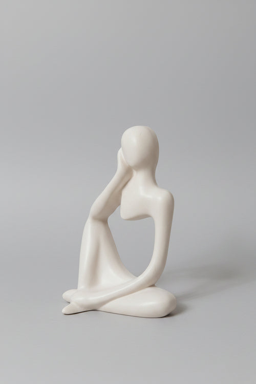 A minimalist white ceramic figurine of a seated, contemplative female figure with a blank face, resting on a light surface.