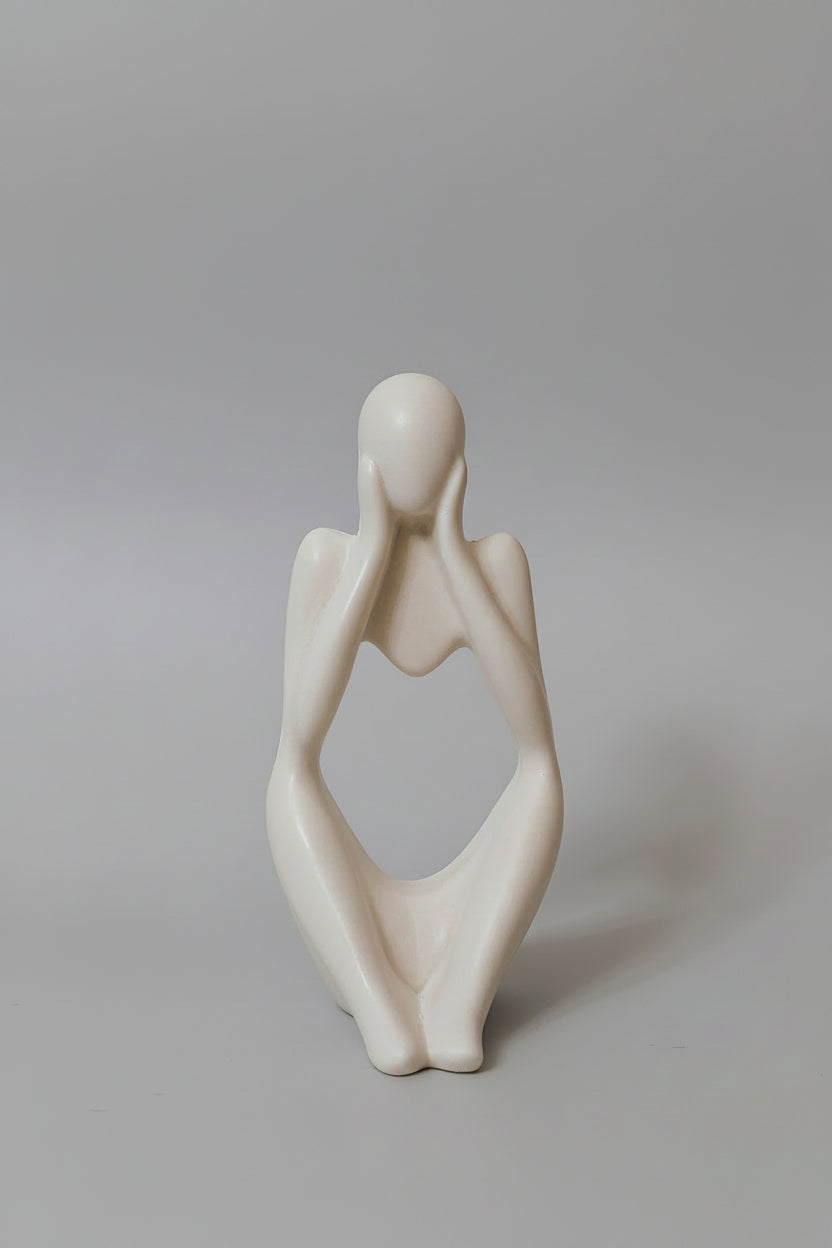 A minimalist white ceramic figurine of a seated figure with hands over face.