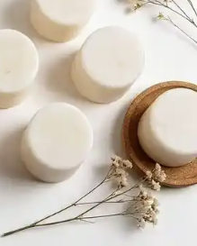 Several round white wax melts are arranged on a white surface, with one bar resting on a small round wooden coaster and dried floral stems placed around them.