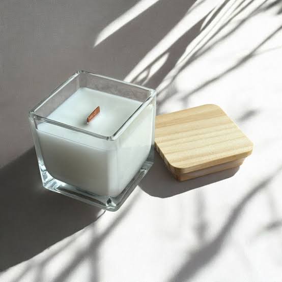 A square glass jar candle with white wax and a small wooden wick, shown next to a light natural wood lid.
