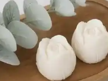 Two white rose-shaped molded wax melts displayed on a wooden tray with decorative eucalyptus leaves.