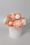 A decorative floral arrangement in a round white box, featuring peach, cream, and white sculpted flowers with dried beige filler, set against a plain beige wall.